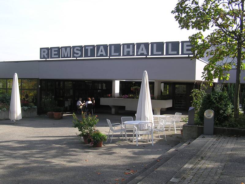 Das Bild zeigt die Remstalhalle mit einer Terrasse, auf der weiße Tische und Stühle sowie Sonnenschirme stehen, und es sind einige Menschen zu sehen.