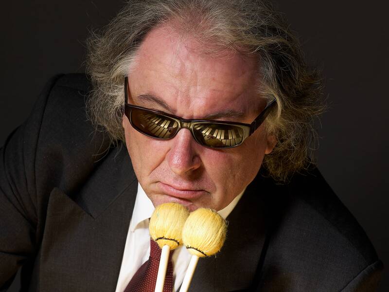 Eine Person mit Sonnenbrille und grauem Haar hält zwei Schlägel vor ihrem Gesicht, während sich im Brillenglas die Tasten eines Klaviers spiegeln.