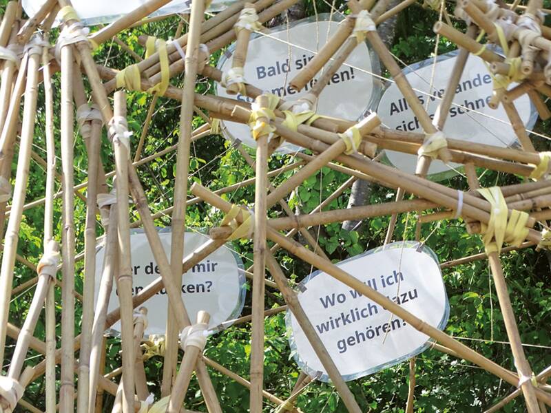 Ein Bambusgerüst im Freien ist mit Schildern versehen, die Fragen wie "Wo will ich wirklich dazugehören?" und andere Fragen zum Nachdenken zeigen.