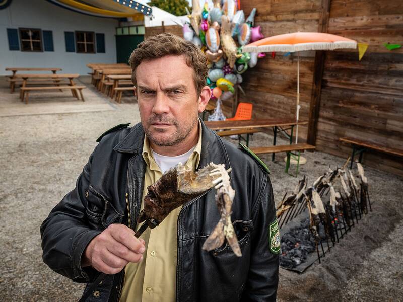 Ein Mann in einer schwarzen Jacke hält eine gegrillte Fischgräte und steht vor einem Holztisch mit Luftballons im Hintergrund.